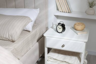 Alarm saati, bir bardak su, hasır kutu ve yatağın yanında kitap olan komodin.