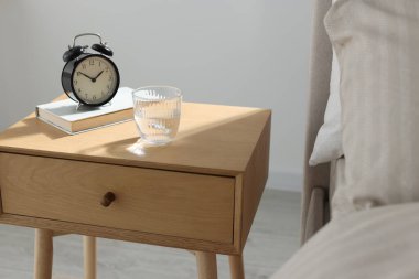 Çalar saati olan komodin, yatağın yanında bir bardak su ve kitap, yakın plan.