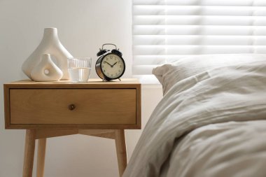 Komodinin yanında çalar saat, bir bardak su ve yatağın yanında vazolar.