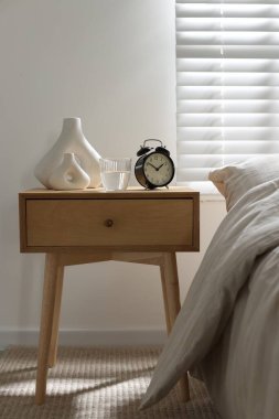 Komodinin yanında çalar saat, bir bardak su ve yatağın yanında vazolar.