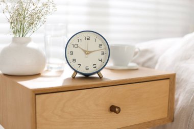 Komodinin yanında çalar saat, vazoda çiçekler, yatağın yanında bir bardak su ve fincan, yakın plan.