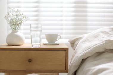 Komodinin yanında bir bardak su, vazoda çiçekler ve yatağın yanında bir fincan.