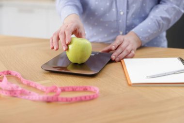 Mutfak terazisinde elmayı tartan kadın, kapalı masa, yakın plan.