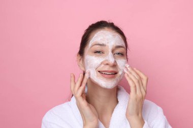 Spa günü. Güzel bir kadın pembe arka planda yüzüne maske takıyor.