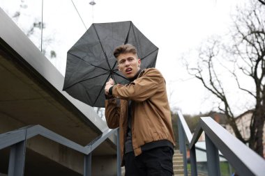 Kırık şemsiyeli bir adam açık havada yağmur altında rüzgarla savaşıyor.