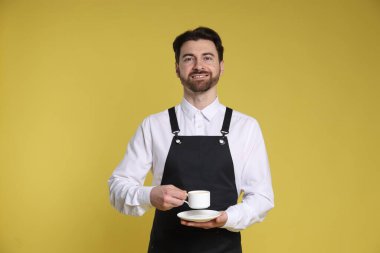 Sarı arka planda bir fincan sıcak içecekle gülümseyen baristanın portresi.