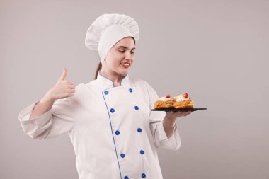 Üniformalı bir şekerci lezzetli profiteroller tutuyor ve açık gri arka planda baş parmaklarını gösteriyor.