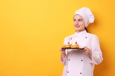 Üniformalı şekerlemeci turuncu arka planda lezzetli profiteroller tutuyor, mesaj için yer var.