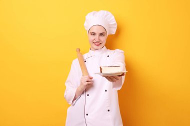 Üniformalı pastacı elinde oklavayla turuncu arka planda lezzetli bir pasta tutuyor.