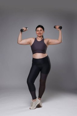 Ayrıca spor elbiseli bir kadın gri arka planda dambıllarla egzersiz yapıyor.