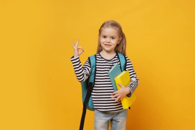 Sırt çantalı ve kitaplı tatlı kız sarı arka planda iyi bir jest gösteriyor.