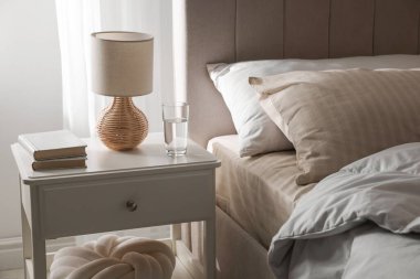 Yatağın yanında kitap, lamba ve bir bardak su olan komodin.