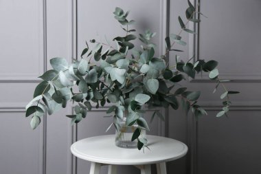 Gri duvarın yanındaki masada vazoda okaliptüs dalları var.