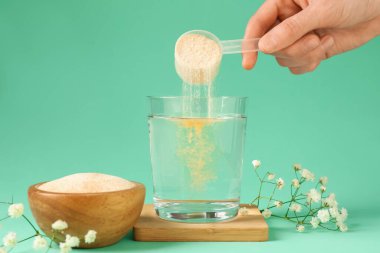 Kadın bardağa kolajen tozu ekliyor. Turkuaz arka planda su var. Yakın plan.