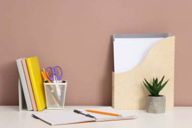 Beyaz ofis masasında koyu bej arka planda farklı kırtasiye ve çiçek dekoru.