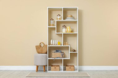 Shelving unit with dishware, jars of different products and other stuff near beige wall indoors