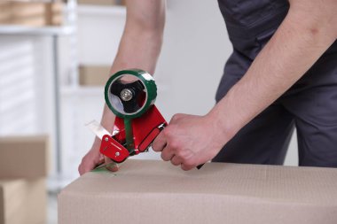 Karton kutuyu kapalı bir yere kapatmak için yapıştırıcılı bant kullanan bir adam.