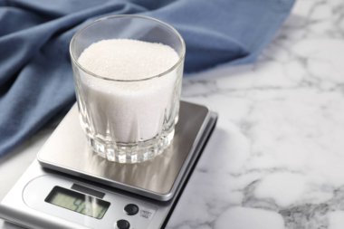 Dijital mutfak terazisi, beyaz mermer masa üzerinde bir bardak şeker, yakın plan.