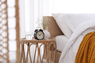 Komodinin yanında çalar saat, vazoda çiçekler, yatağın yanında bir bardak su ve bardaklar.