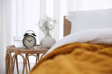 Komodinin yanında çalar saat, vazoda çiçekler, yatağın yanında bir bardak su ve bardaklar.