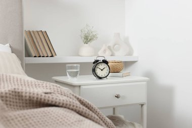 Alarm saati, bir bardak su, hasır kutu ve yatağın yanında kitap olan komodin.