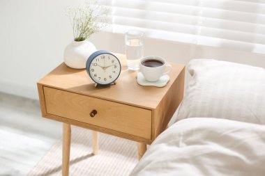 Çalar saati olan komodin, vazoda çiçekler, yatağın yanında bir bardak su ve kahve.