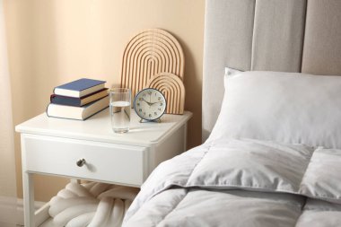 Yatağın yanında kitap, dekor, bir bardak su ve çalar saat olan komodin.