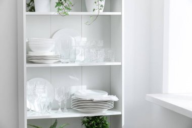 Beyaz duvarın yanındaki raflarda temiz bardaklar, tabaklar ve yeşil bitkiler var.