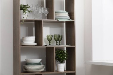 Beyaz duvarın yanındaki raflarda temiz bardaklar, tabaklar ve yeşil bitkiler var.