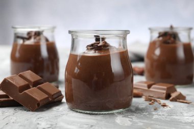 Cam kavanozlarda lezzetli puding ve açık gri masada çikolata parçaları, yakın plan.