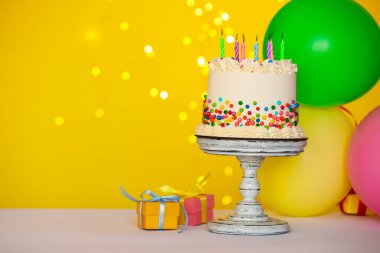 Rengarenk serpiştirilmiş lezzetli bento keki, yanan mumlar ve masanın üzerinde sarı arka plana karşı diğer şenlikli dekorlar, yazı için yer. Bokeh etkisi