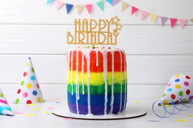 Lezzetli gökkuşağı doğum günü pastası, beyaz ahşap arka planda parti şapkaları ve konfeti.