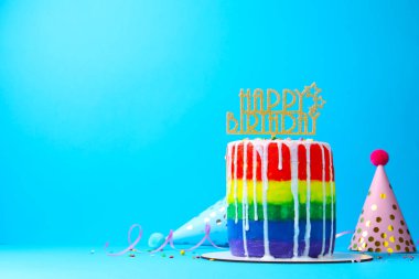 Lezzetli gökkuşağı doğum günü pastası, süslemeler ve açık mavi arka planda parti şapkaları, metin için yer