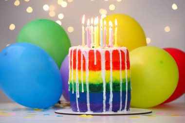 Lezzetli gökkuşağı doğum günü pastası, yanan mumlar, renkli balonlar ve beyaz masanın üzerindeki konfetiler, gri arka planda bulanık ışıklar, yakın plan