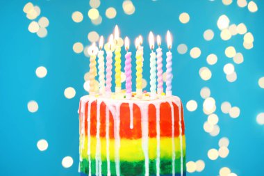 Enfes gökkuşağı doğum günü pastası açık mavi arka planda yanan mumlar, bulanık ışıklar, yakın plan