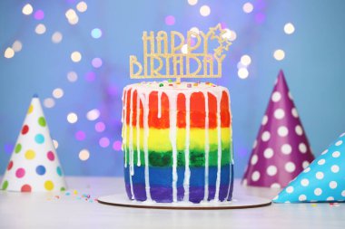 Beyaz masadaki parti şapkalarıyla birlikte nefis gökkuşağı pastası açık mavi arka plan, bulanık ışıklar, yakın plan