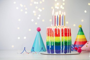 Enfes gökkuşağı doğum günü pastası, yanan mumlar, flamalar, serpiştirmeler ve parti şapkaları beyaz masanın üzerinde bulanık ışıklı arka planda, yakın plan. Metin için boşluk