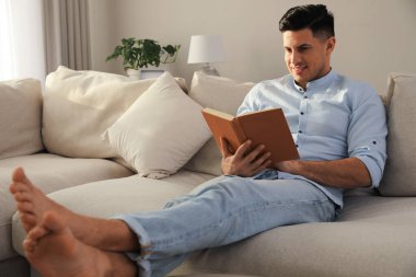 Oturma odasındaki kanepede kitap okuyan adam.