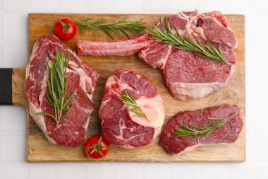 Pieces of raw beef, rosemary and tomatoes on white tiled table, top view
