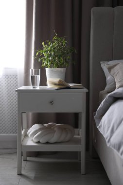 Yatağın yanında kitap, çiçek ve bir bardak su olan komodin.