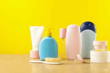 Bebek kozmetik ürünleri ve banyo aksesuarları ahşap masada, sarı arka planda. Metin için boşluk