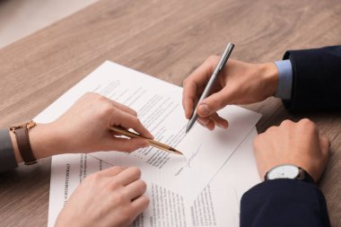 Kadın belgeye işaret ediyor ve adam ahşap masaya imza atıyor, yakın plan.