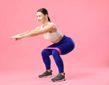 Spor kıyafetli bir kadın pembe arka planda elastik bantla egzersiz yapıyor.