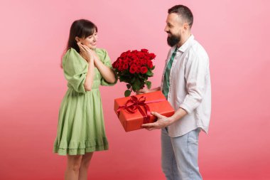 Kırmızı gül buketini ve hediye kutusunu pembe arka plandaki sevgilisine sunan bir adam.