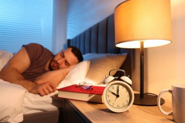 Evde uyuyan genç adam, çalar saate, kitaba ve gözlüğe odaklan.