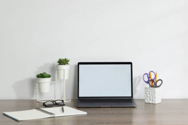 Laptop, farklı kırtasiye malzemeleri, gözlük ve çiçek dekoru beyaz arka planda ahşap ofis masasında.