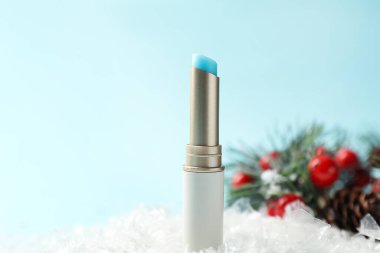 Nemlendirici dudak kremi ve yapay kar üzerindeki Noel süsü açık mavi arka planda, yakın plan. Kış bakımı