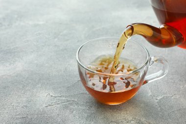 Gri masada çaydanlıktan fincana taze demlenmiş çay dökülüyor, yakın plan. Metin için boşluk