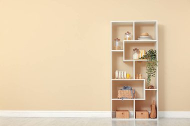 Shelving unit with dishware, jars of different products and other stuff near beige wall indoors, space for text