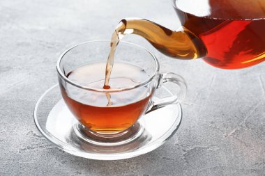 Gri masada çaydanlıktan fincana taze demlenmiş çay dökülüyor, yakın plan.